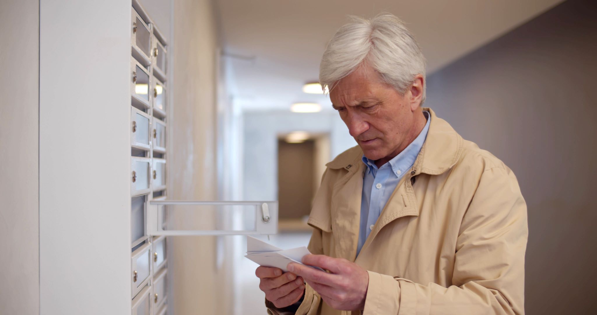 Senior man stands at indoor mailbox in foyer looking confused.